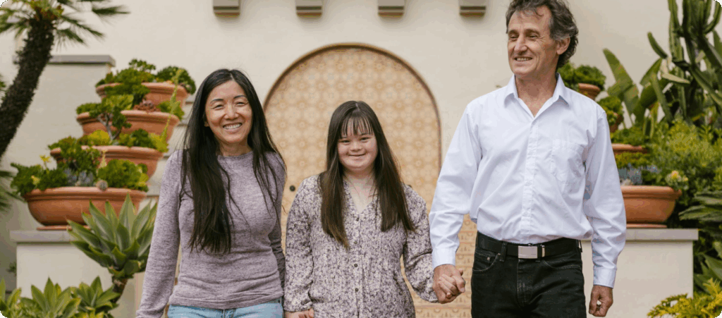 A young girl with Down syndrome holding hands with a man and a woman on her left and right sides.