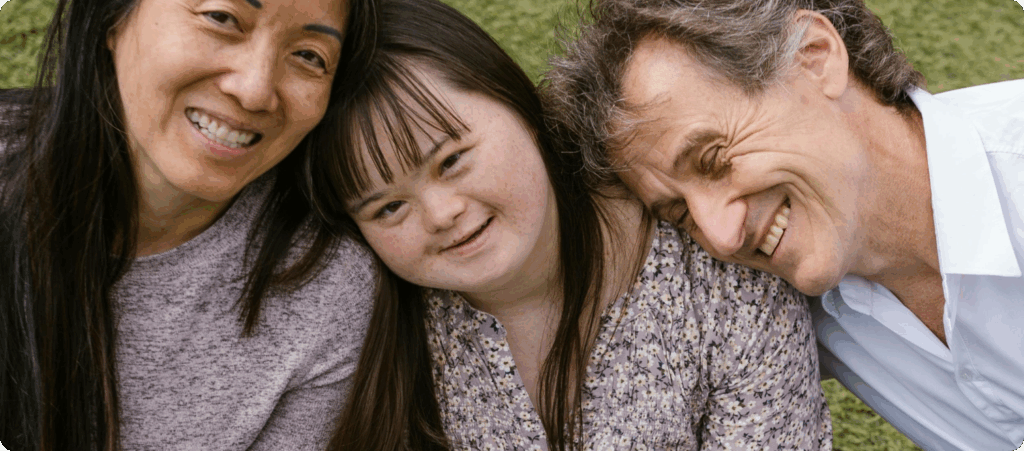A young girl with Down syndrome with a man and woman leaning gently on her, all smiling.