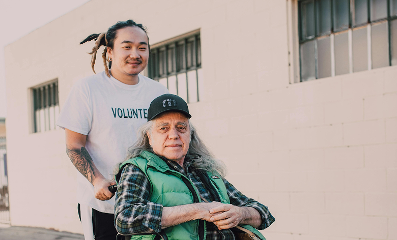 A young man smiling as he pushes an elderly man in a wheelchair, both looking at the camera.