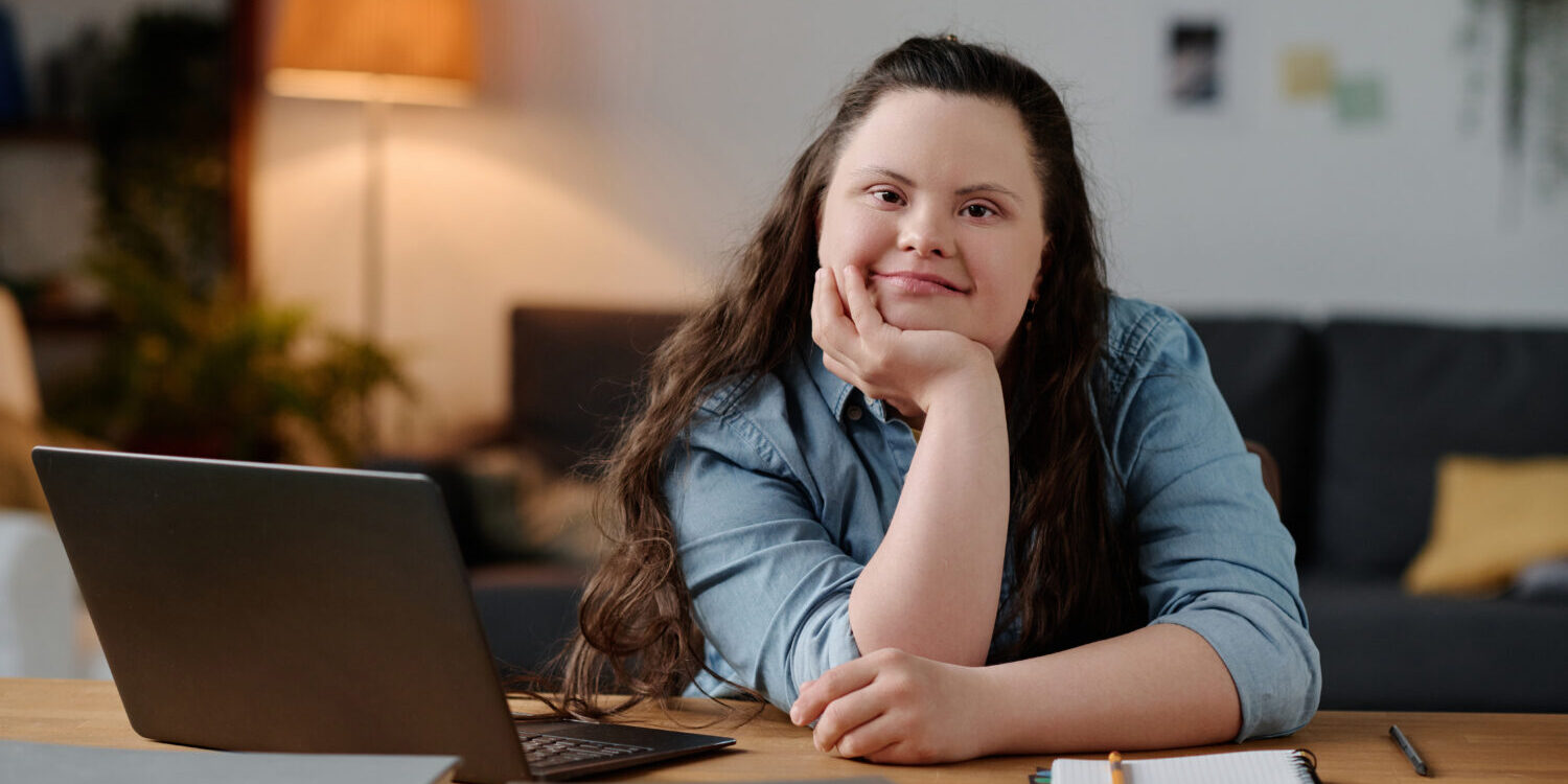 Girl with down syndrome studying at home Portrait of girl with down syndrome looking at camera while sitting at desk with laptop in the living room