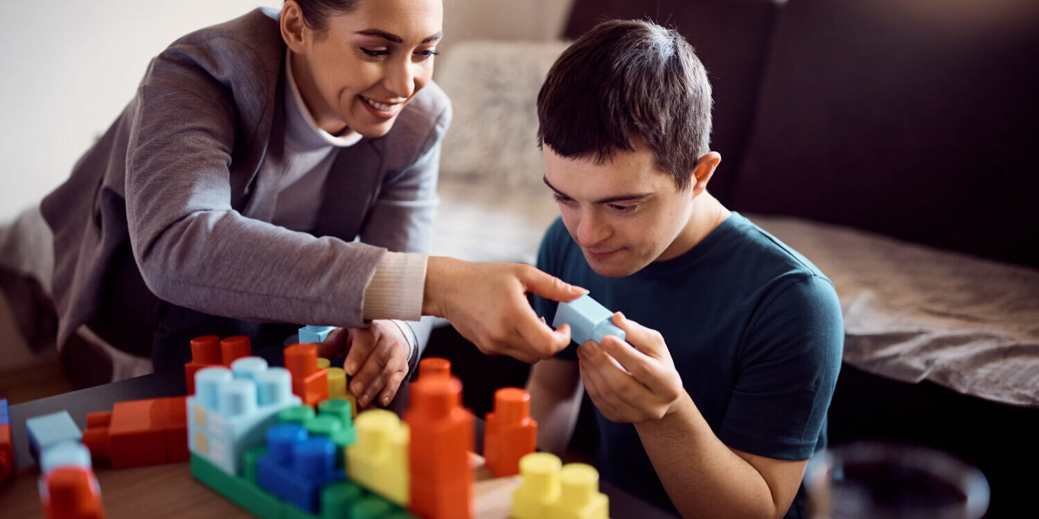 Man with down syndrome enjoying while stacking toy blocks with his psychologist at home. Happy psychologist and man who has down syndrome playing with toy blocks during home visit.